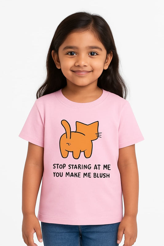 A young Indian girl with short dark hair stands smiling in a pale yellow T-shirt featuring a cartoon orange cat from behind and the text “STOP STARING AT ME YOU MAKE ME BLUSH.” He pairs the shirt with blue jeans and stands against a simple, light-colored background suitable for an e-commerce catalog.