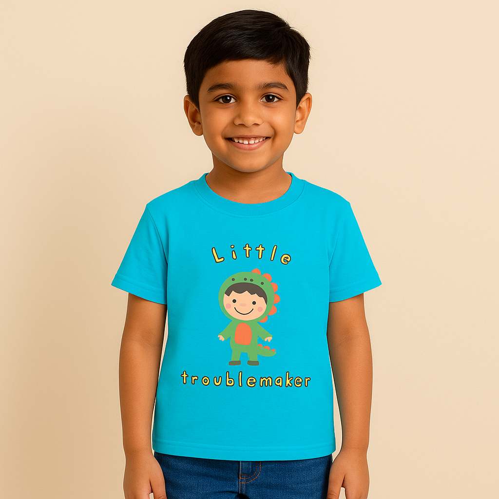 A digital photograph of a young Indian boy, around 8 years old, with fair skin tone, smiling and standing naturally. He is wearing a bright blue T-shirt with a cute “Little Troublemaker” design showing a cartoon child dressed in a green dinosaur costume. The look is completed with casual denim jeans, posed against a clean, neutral studio background with soft lighting, styled for an e-commerce clothing catalog.