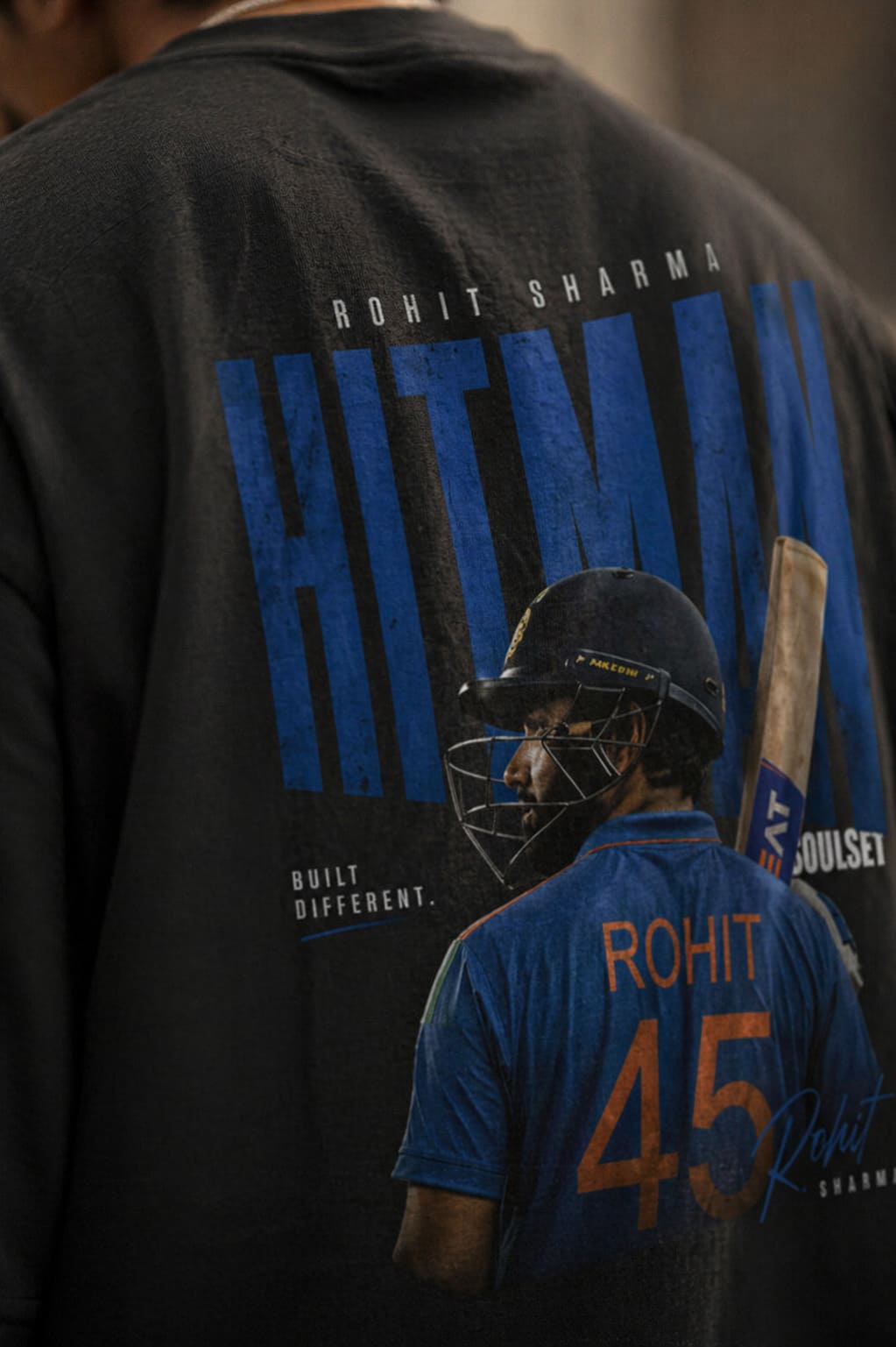 Person wearing a black shirt with a graphic of a cricket player and 'Rohit Sharma' text, standing in front of a blurred background.