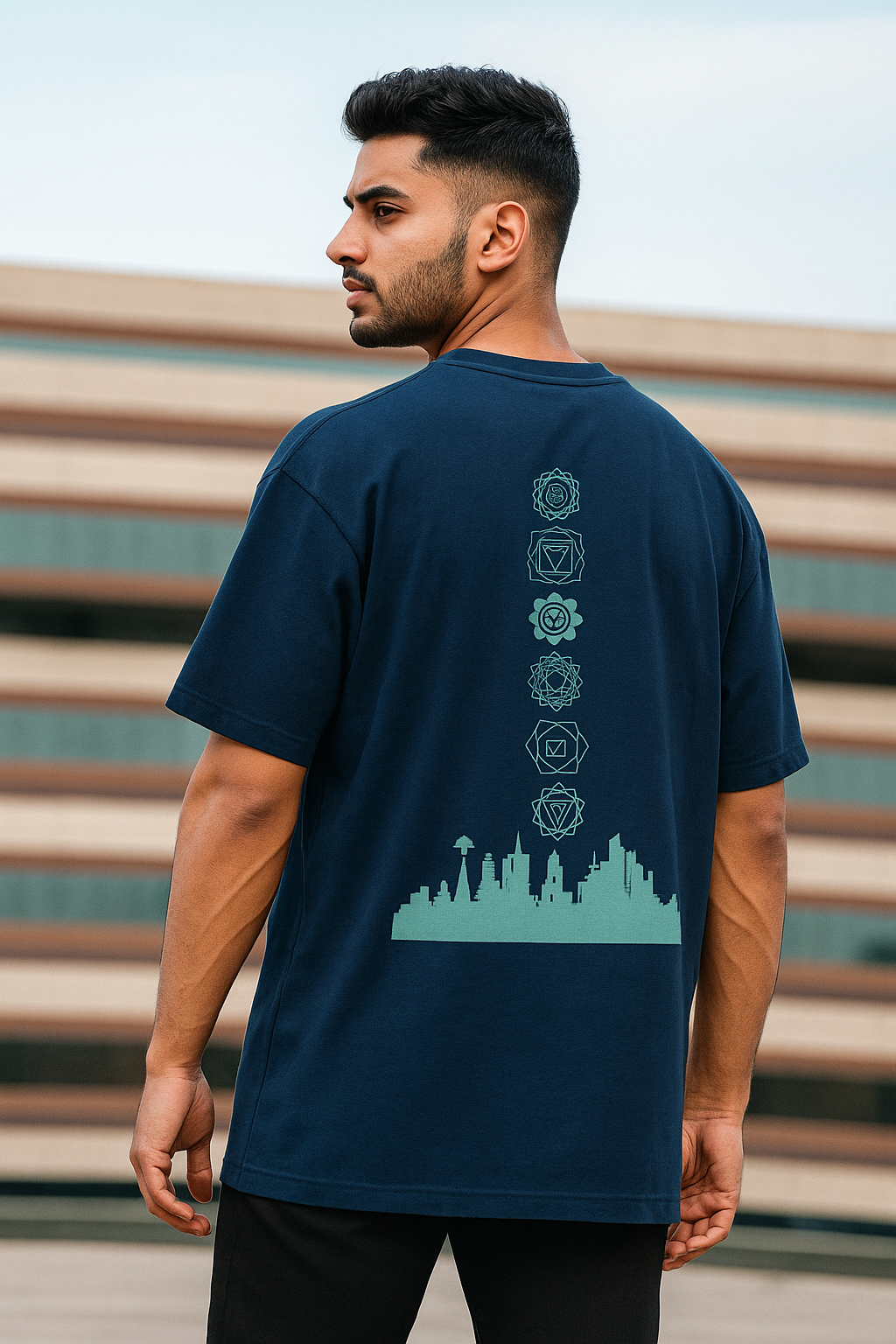 Indian male model wearing a navy blue oversized t-shirt with a bold back graphic featuring chakra symbols above a city skyline. The fit is modern and relaxed, with the model standing confidently facing the camera, showcasing the detailed back design. Perfect for streetwear and urban fashion.