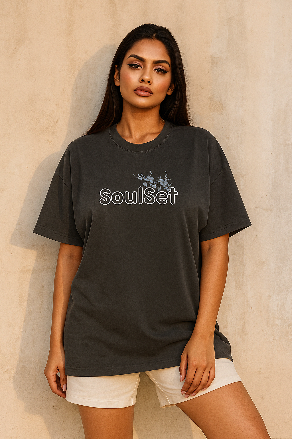 A confident young Indian woman with long dark hair wears an oversized black SoulSet T-shirt featuring a floral design on the front, standing against a textured beige concrete wall in warm natural ligh