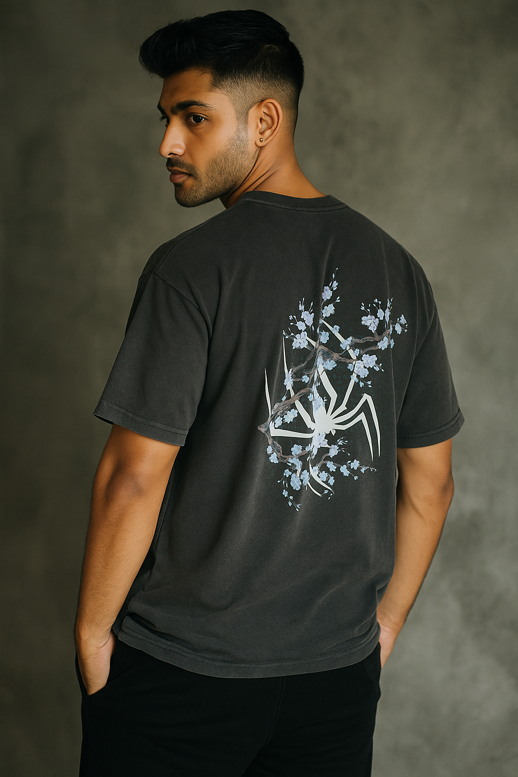 Confident young South Asian male model wearing a black oversized T-shirt, showcasing the back design with a white spider and blue floral branches, standing stylishly in a relaxed pose.