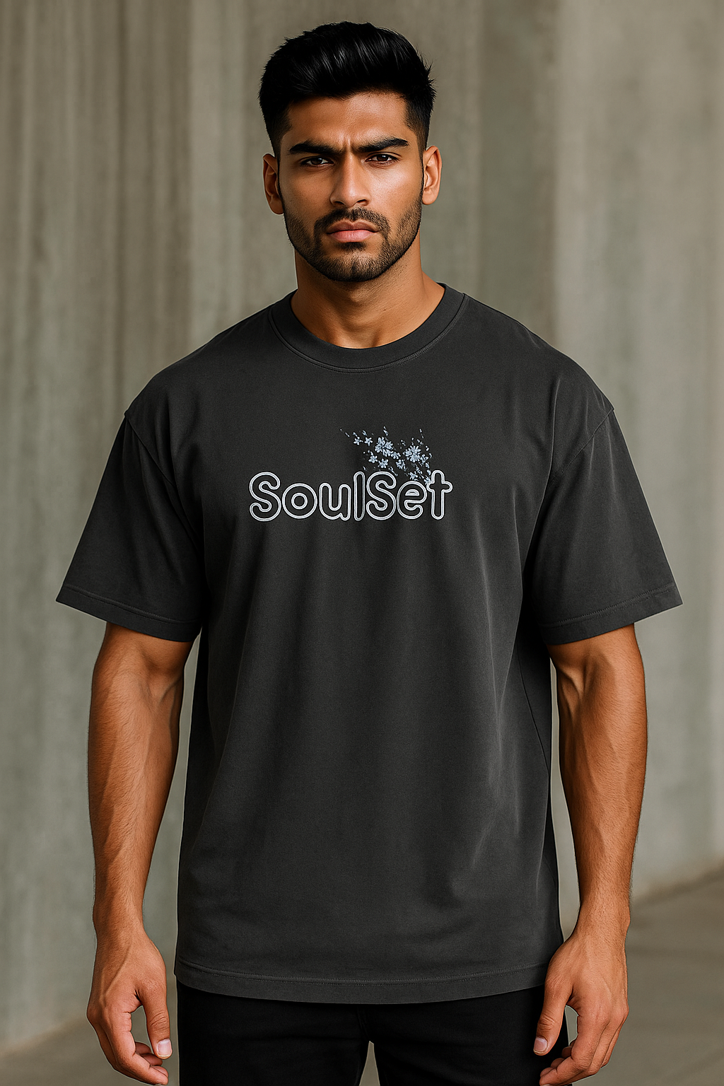 A confident young Indian man with a fit build and neatly styled dark hair wears an oversized black SoulSet T-shirt with a floral front design, standing against a gray concrete wall in soft natural light