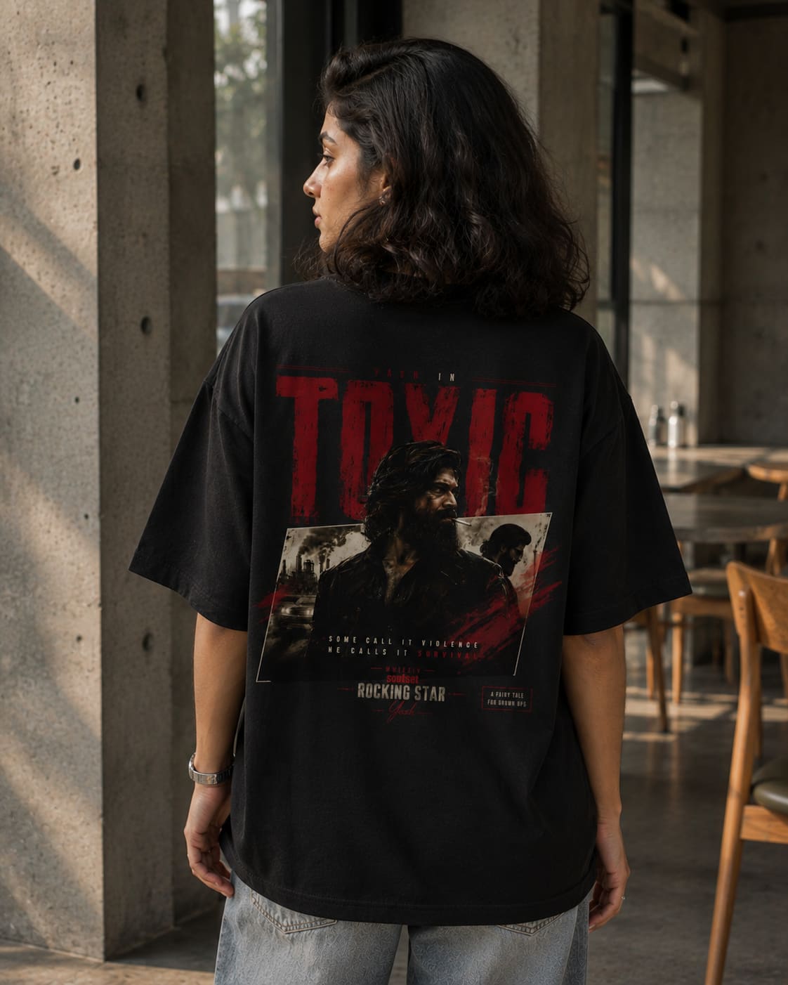A woman wearing an oversized black graphic T-shirt with a large red and white print on the back, standing in a modern concrete-walled indoor space.