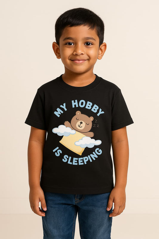 A cheerful young Indian boy with short dark hair smiles brightly in a studio with a neutral beige background. She is dressed in a red T-shirt featuring a sleeping bear graphic and the text “MY HOBBY IS SLEEPING,” along with casual blue jeans.