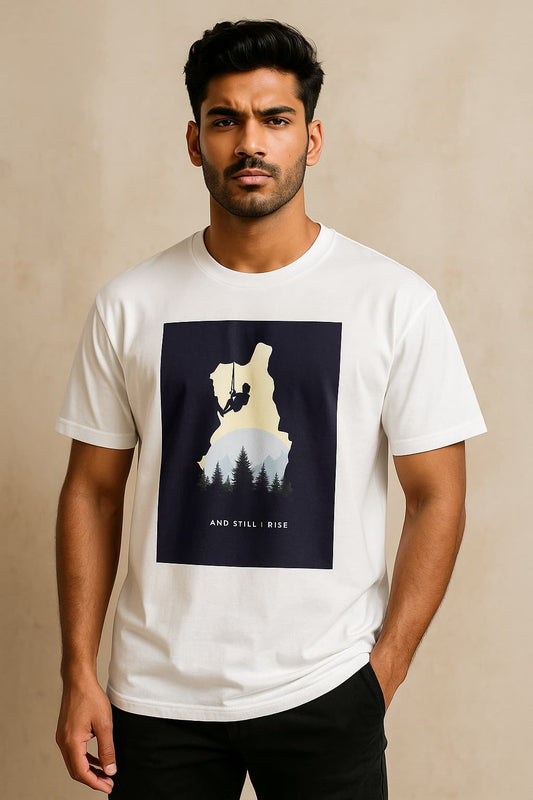 A young South Asian male model stands confidently wearing a yellow oversized T-shirt with a bold adventure-themed print showing a climber silhouette, forest, and sun with the words “And Still I Rise.” He poses stylishly against an aesthetic background for an e-commerce catalog.