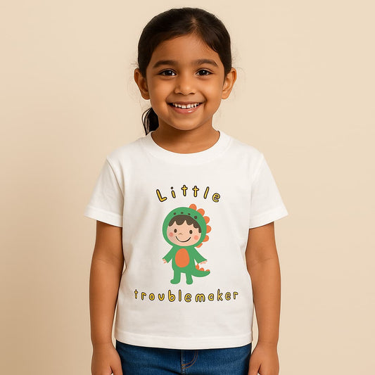 A digital photograph of a young Indian girl, around 8 years old, with light skin tone, smiling and standing naturally. She is wearing a white T-shirt with a playful “Little Troublemaker” design featuring a cartoon child in a green dinosaur costume. The outfit is paired with casual denim jeans, and the scene is set against a clean, neutral studio background with soft lighting, styled for an e-commerce clothing catalog.