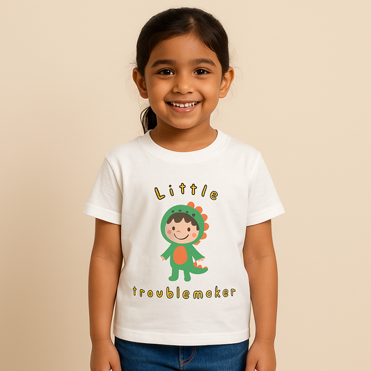 A digital photograph of a young Indian girl, around 8 years old, with light skin tone, smiling and standing naturally. She is wearing a white T-shirt with a playful “Little Troublemaker” design featuring a cartoon child in a green dinosaur costume. The outfit is paired with casual denim jeans, and the scene is set against a clean, neutral studio background with soft lighting, styled for an e-commerce clothing catalog.