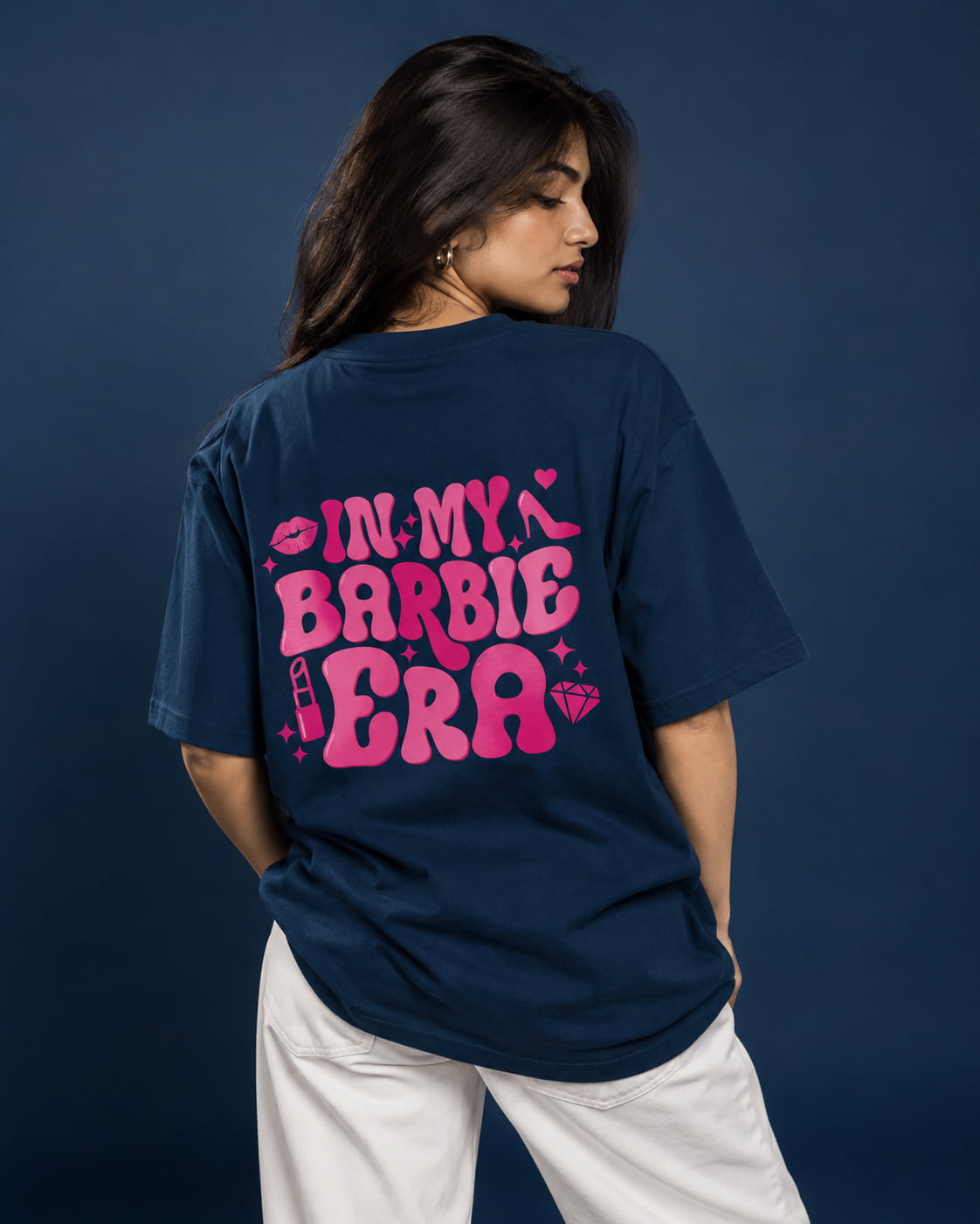 A woman wearing a navy blue oversized T-shirt with a bright pink graphic that reads 'IN MY BARBIE ERA' on the back, paired with white pants, posed against a dark blue studio background