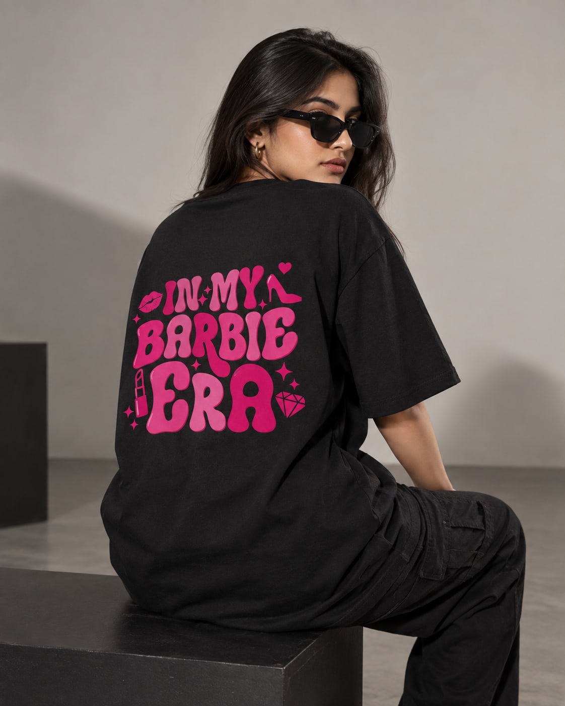 A woman wearing a black oversized T-shirt and black cargo pants sits on a dark block, shown from the back. The shirt features a large bright pink graphic reading 'IN MY BARBIE ERA' with small decorative icons.
