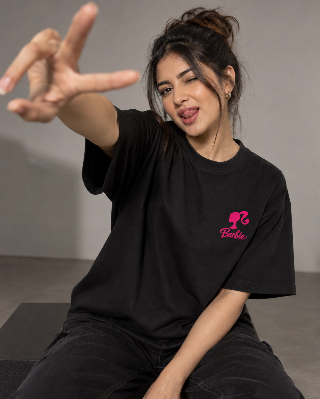 A woman wearing a black t-shirt with a pink Barbie logo on the left chest, posing with one hand extended in a gesture. The background is a clean, neutral gray studio setting.