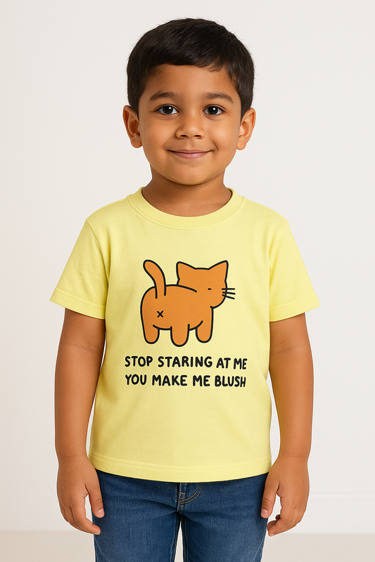 A young Indian boy with short dark hair stands smiling in a pale yellow T-shirt featuring a cartoon orange cat from behind and the text “STOP STARING AT ME YOU MAKE ME BLUSH.” He pairs the shirt with blue jeans and stands against a simple, light-colored background suitable for an e-commerce catalog.