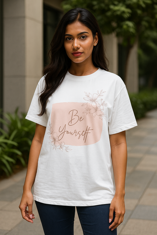 nfident young Indian woman in her 20s wearing a white oversized T-shirt with ‘Be Yourself’ text and floral design, posing outdoors in a modern urban background for an e-commerce fashion photoshoot
