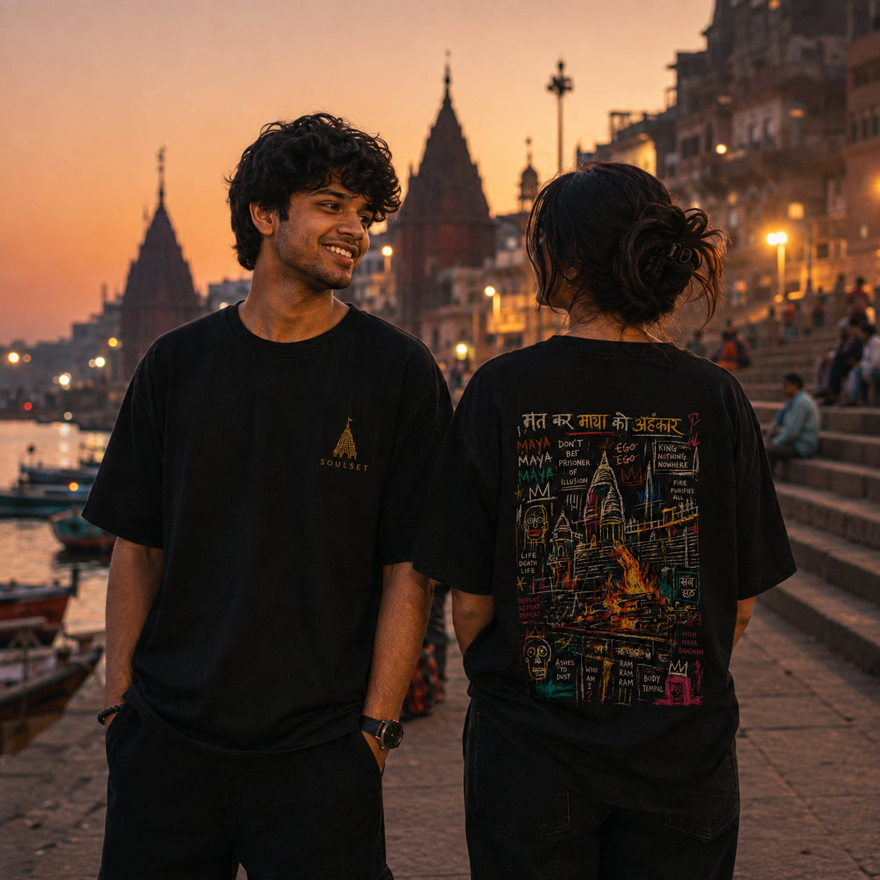 Two people standing on a riverside walkway at sunset, wearing matching black graphic T-shirts. The person on the left faces forward with a small gold chest logo, and the person on the right faces away showing a large multicolor back graphic with text and cityscape artwork.