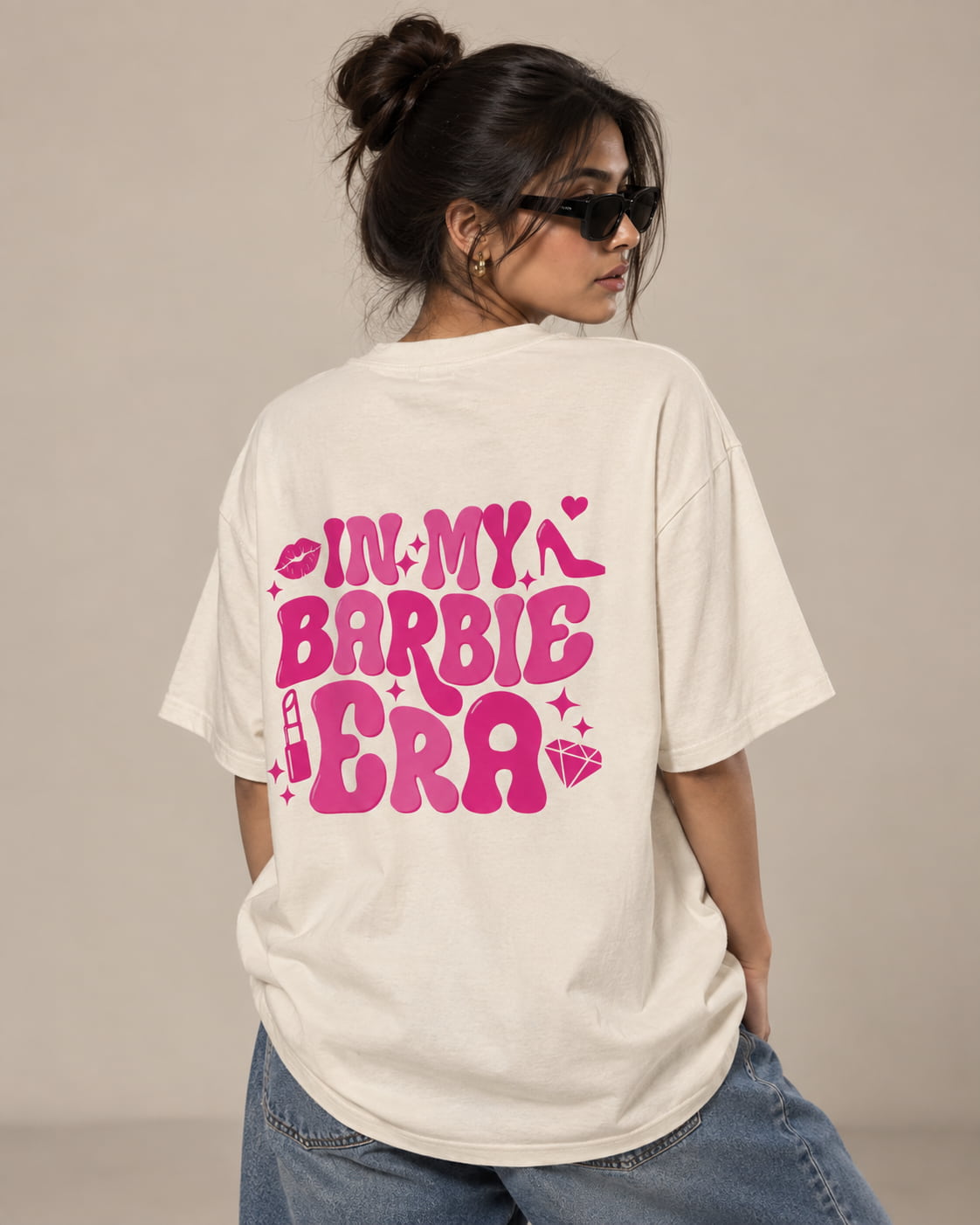 A woman wearing an oversized cream-colored t-shirt with a vibrant pink graphic on the back that reads 'IN MY BARBIE ERA' with small pink hearts, lollipops, and a diamond, paired with blue jeans against a neutral beige background.