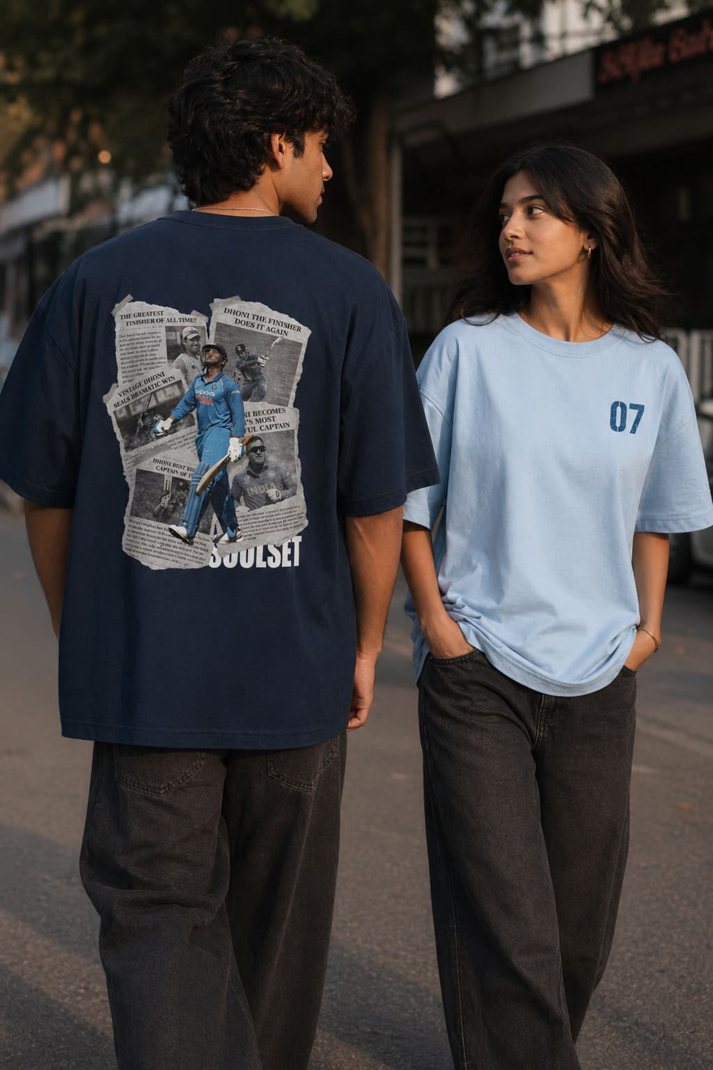 Two people standing on a street, one wearing a navy t-shirt with a graphic design and the other wearing a light blue t-shirt with '07' printed on it.