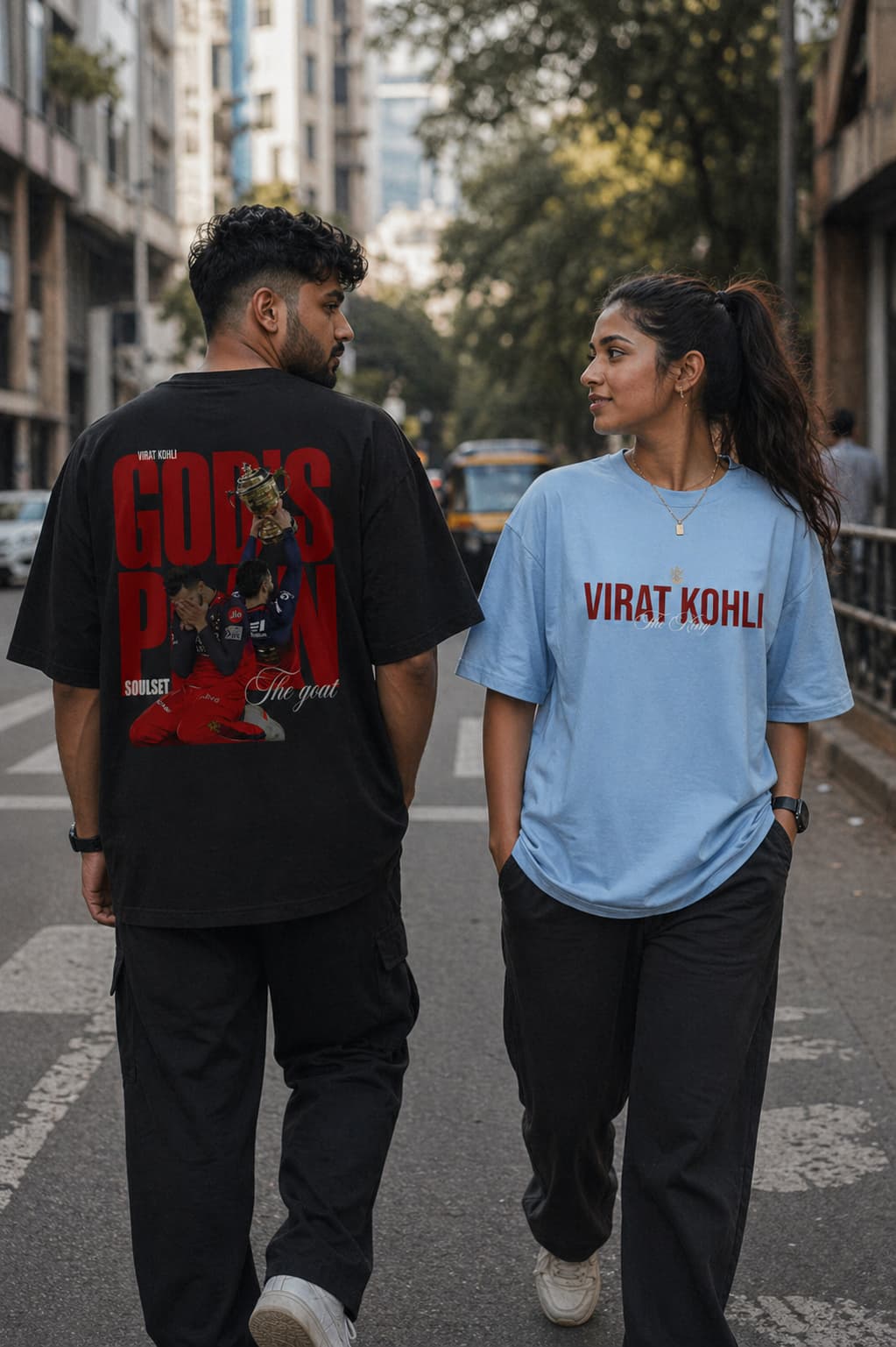 Two people walking on a street, one wearing a black t-shirt with red graphics and text, the other wearing a blue t-shirt with 'Virat Kohli' text.