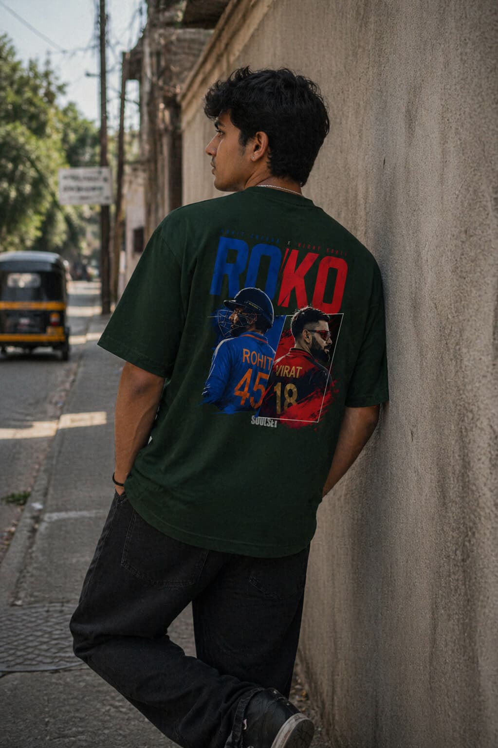 Person wearing a green t-shirt with a colorful graphic design leaning against a wall on a street.