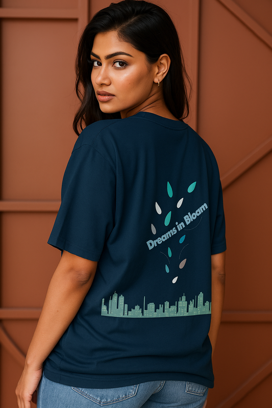 Woman wearing a navy blue t-shirt with a city skyline and 'Dreams in Bloom' text on the back against a brown wall.
