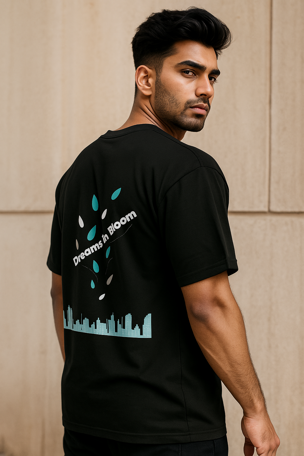 Man wearing a black t-shirt with a logo and text on the back, standing against a beige wall.