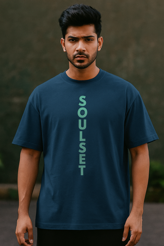 A confident and stylish Indian male model with a fair complexion and a fit build is wearing a navy blue oversized SoulSet t-shirt. The t-shirt features a bold vertical "SOULSET" text design printed in mint green on the front. The model is facing the camera in a relaxed pose, showcasing the front design clearly. The background is modern and minimal, keeping the focus on the t-shirt.
