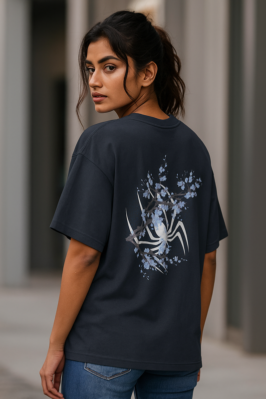 Confident young South Asian female model wearing a black oversized T-shirt, showcasing the back design with a white spider and blue floral branches, standing stylishly in a relaxed pose.