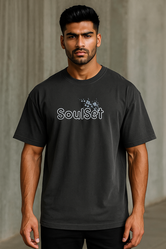 A confident young Indian man with a fit build and neatly styled dark hair wears an oversized black SoulSet T-shirt with a floral front design, standing against a gray concrete wall in soft natural light