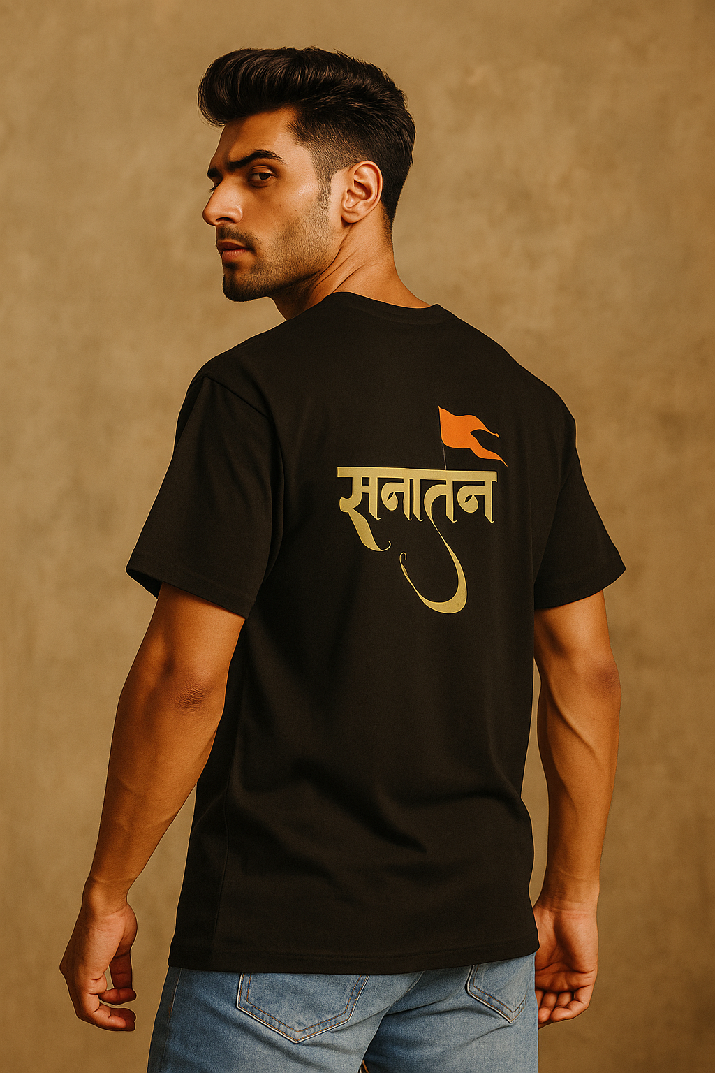 A confident young South Asian male model with fair skin and a fit build wearing a black oversized T-shirt with a bold 'Sanatan' text and saffron flag design on the back. He is posing against a beige textured background, looking over his shoulder with a stylish and bold expression