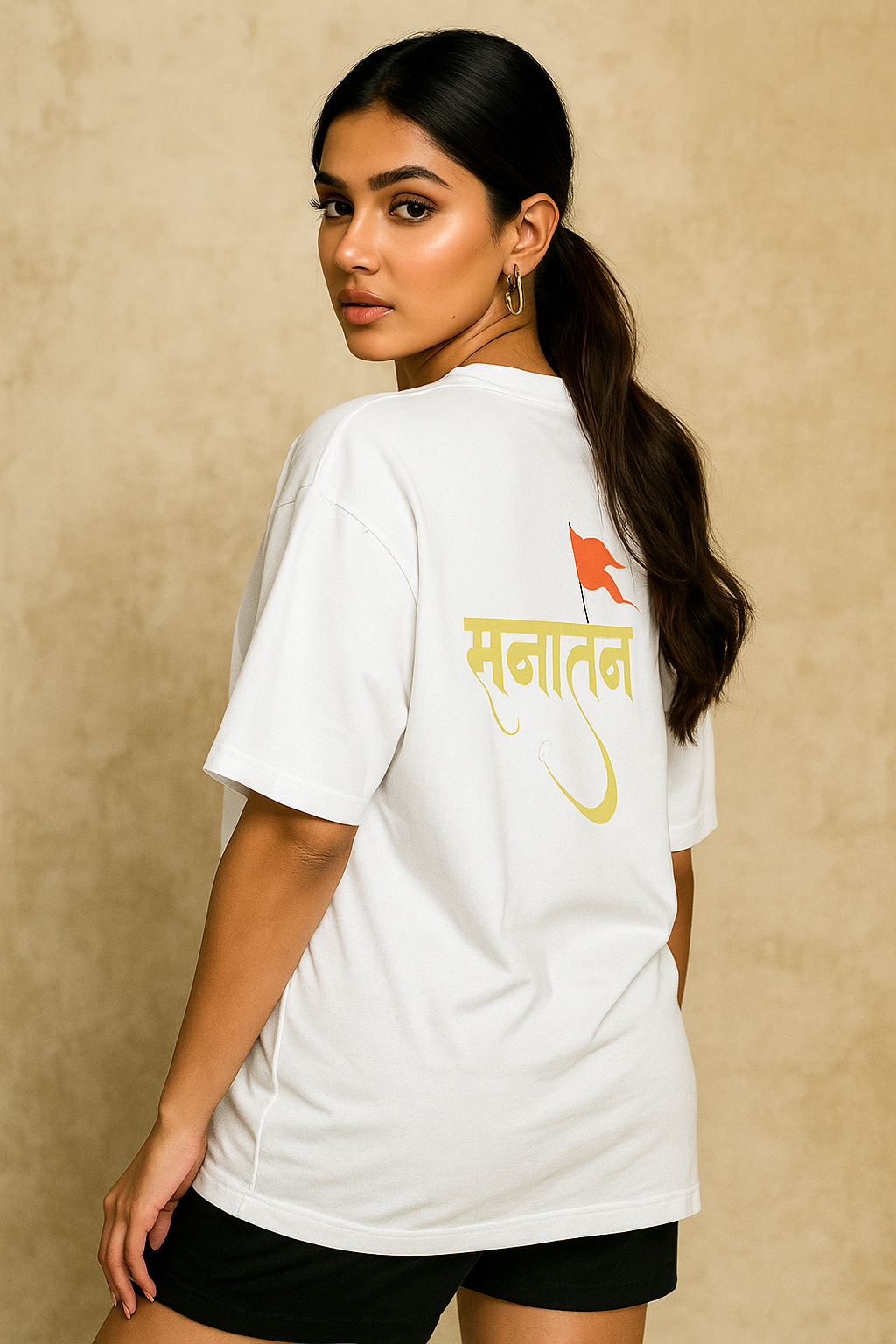 A confident young South Asian female model with fair skin and a fit build wearing a white oversized T-shirt featuring a bold 'Sanatan' text in Devanagari script with a saffron flag design on the back. She poses stylishly against a beige textured background, looking over her shoulder with a bold and elegant expression