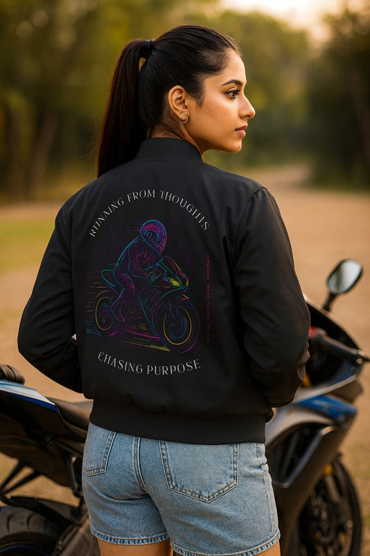 Woman wearing a black jacket with a colorful graphic and text, standing next to a motorcycle in a natural setting.