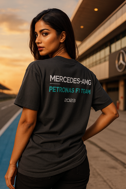A confident Indian female model with fair skin and a fit physique stands in a relaxed pose, facing away to showcase the back of her black oversized Mercedes-AMG Petronas F1 Team t-shirt. The t-shirt features bold white and teal typography with “MERCEDES-AMG,” “PETRONAS F1 TEAM,” and “2023” printed on it. The model is positioned on a Formula 1 pit lane at sunset, with a sleek Mercedes-Benz garage in the background, exuding a bold and stylish F1-themed atmosphere.
