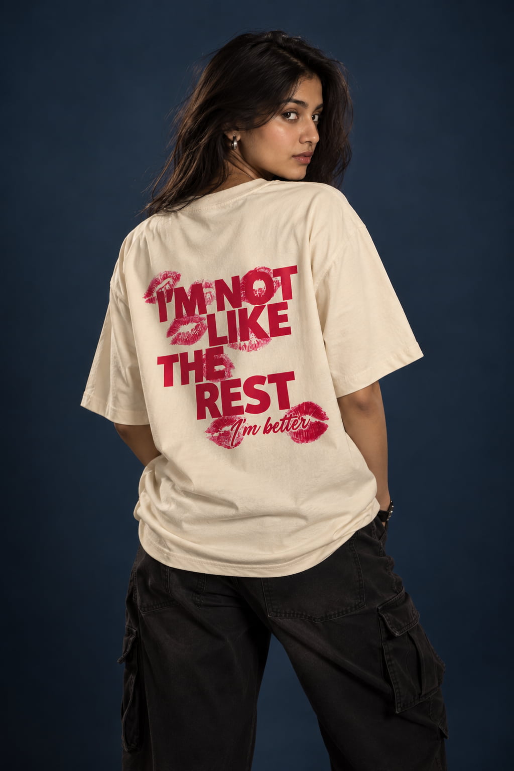 A woman wearing an oversized cream-colored t-shirt with a red graphic text print on the back, paired with black cargo pants, posed against a dark blue studio background.