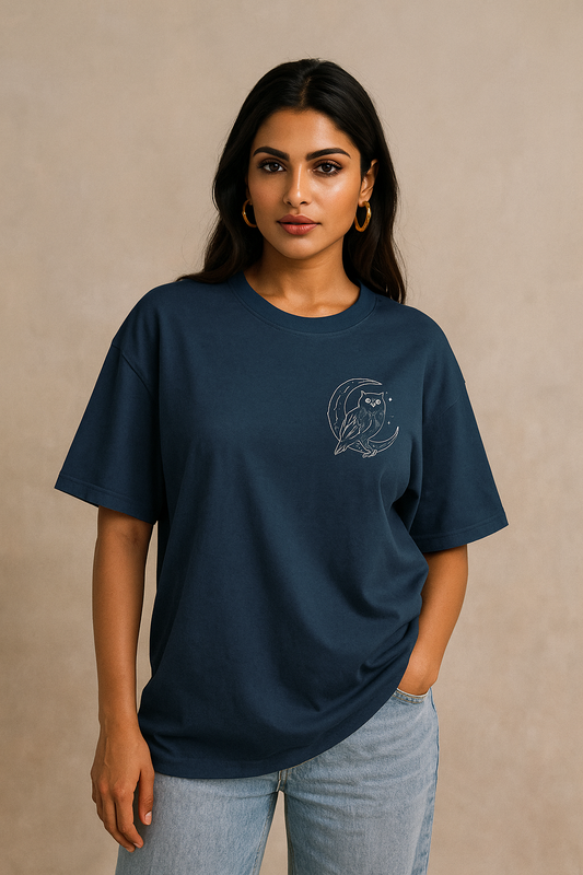 Oversized unisex t-shirt featuring a majestic owl with glowing eyes perched on a crescent moon, surrounded by sacred geometry and stardust detailing on the front. The back showcases an owl in flight fading into mist, with mandala feather rings and the phrase ‘Wisdom Sees All’ above.