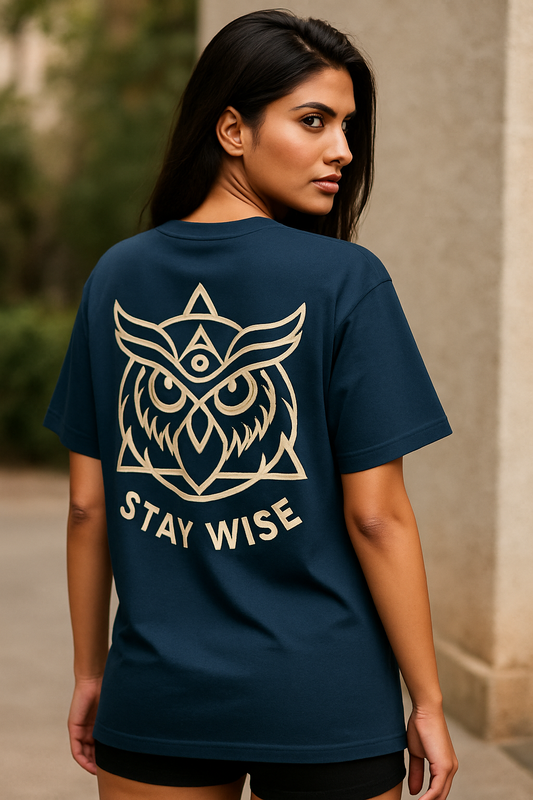 Oversized unisex t-shirt featuring a majestic owl with glowing eyes perched on a crescent moon, surrounded by sacred geometry and stardust detailing on the front. The back showcases an owl in flight fading into mist, with mandala feather rings and the phrase ‘Wisdom Sees All’ above.