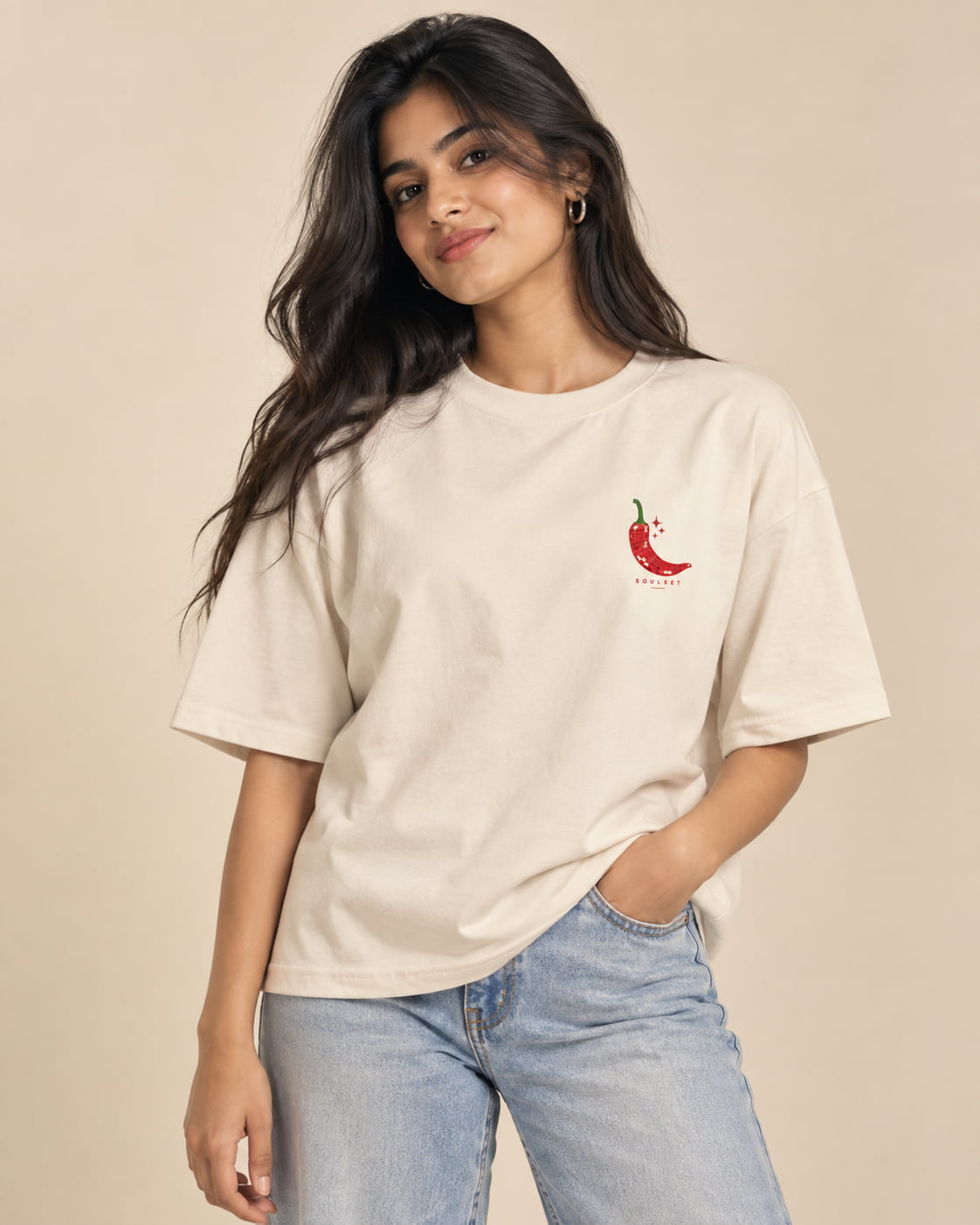 A woman wearing a cream-colored oversized t-shirt with a small red chili pepper graphic on the left chest, paired with light-wash blue jeans, set against a solid beige background.
