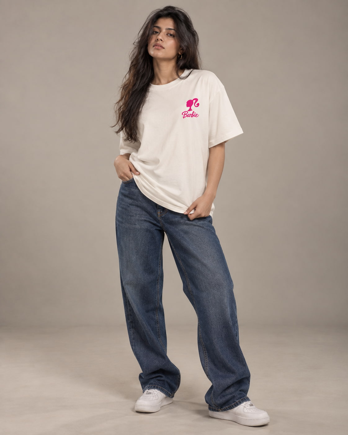 A woman modeling an oversized cream-colored t-shirt with a pink Barbie logo on the chest, paired with wide-leg blue jeans and white sneakers, set against a plain beige studio background.