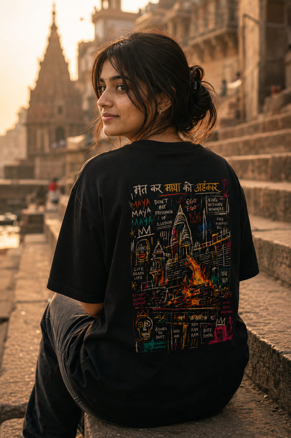 A woman sitting on stone steps outdoors, wearing a black oversized T-shirt with a large colorful graphic print on the back. The background shows a warm, blurred urban architectural scene at sunset.