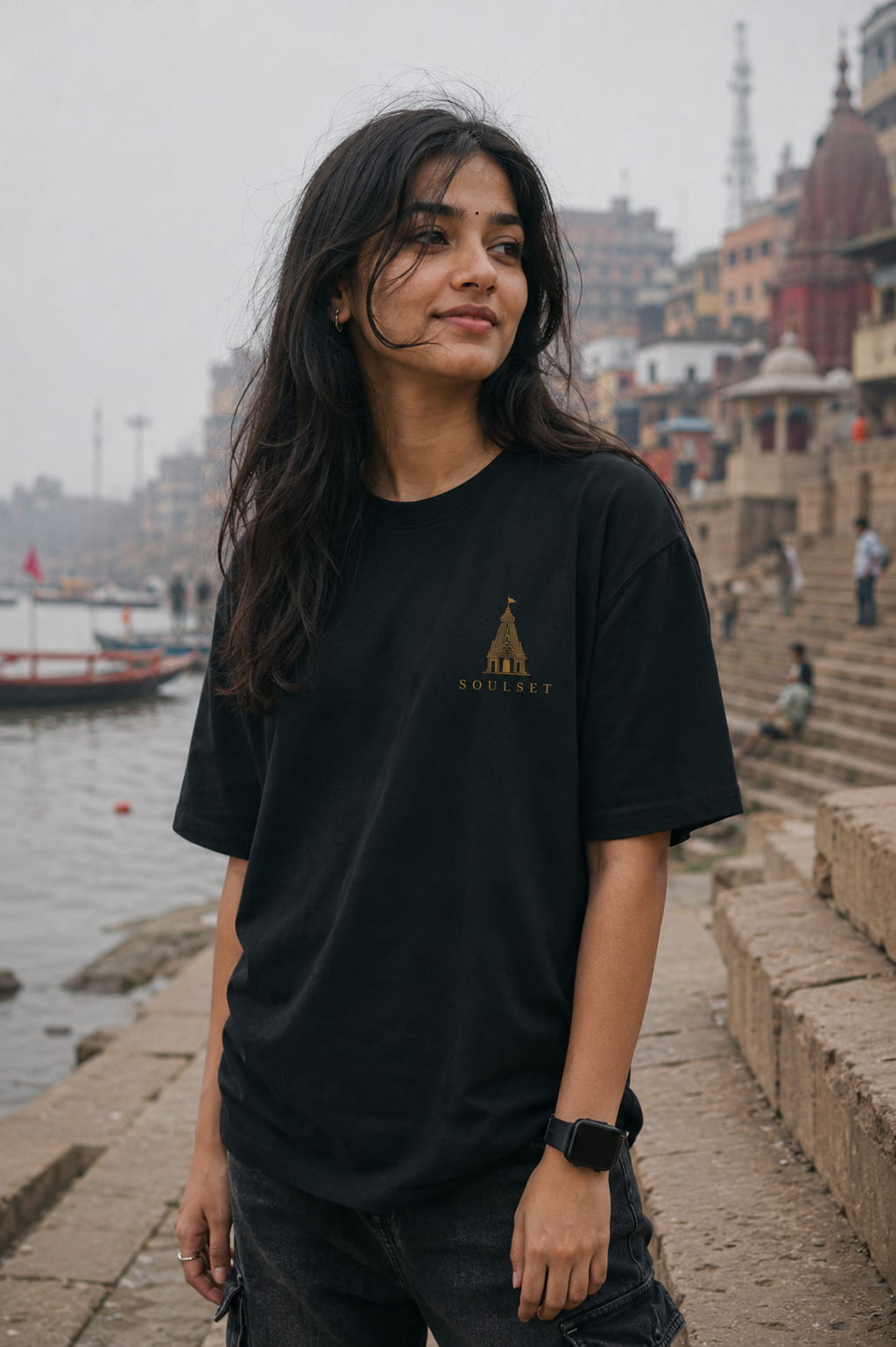 A woman wearing a black t-shirt with a gold temple graphic and the word 'SOULSET' printed on the chest, standing on stone steps by a river with a hazy cityscape in the background.