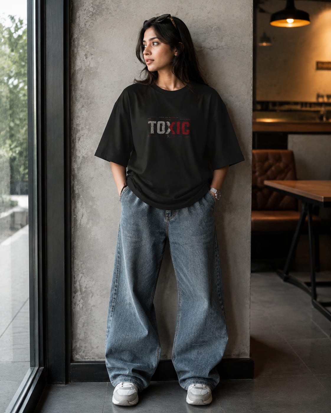 A woman stands indoors leaning against a concrete pillar, wearing an oversized black graphic T-shirt with the word 'TOXIC' printed across the chest in white and red, paired with loose light-wash jeans and white sneakers.