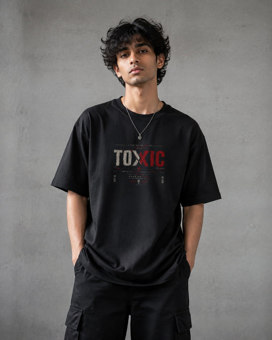 A man wearing a black graphic t-shirt with 'TOXIC' printed on the chest in white and red, paired with black cargo pants, standing against a textured gray wall.