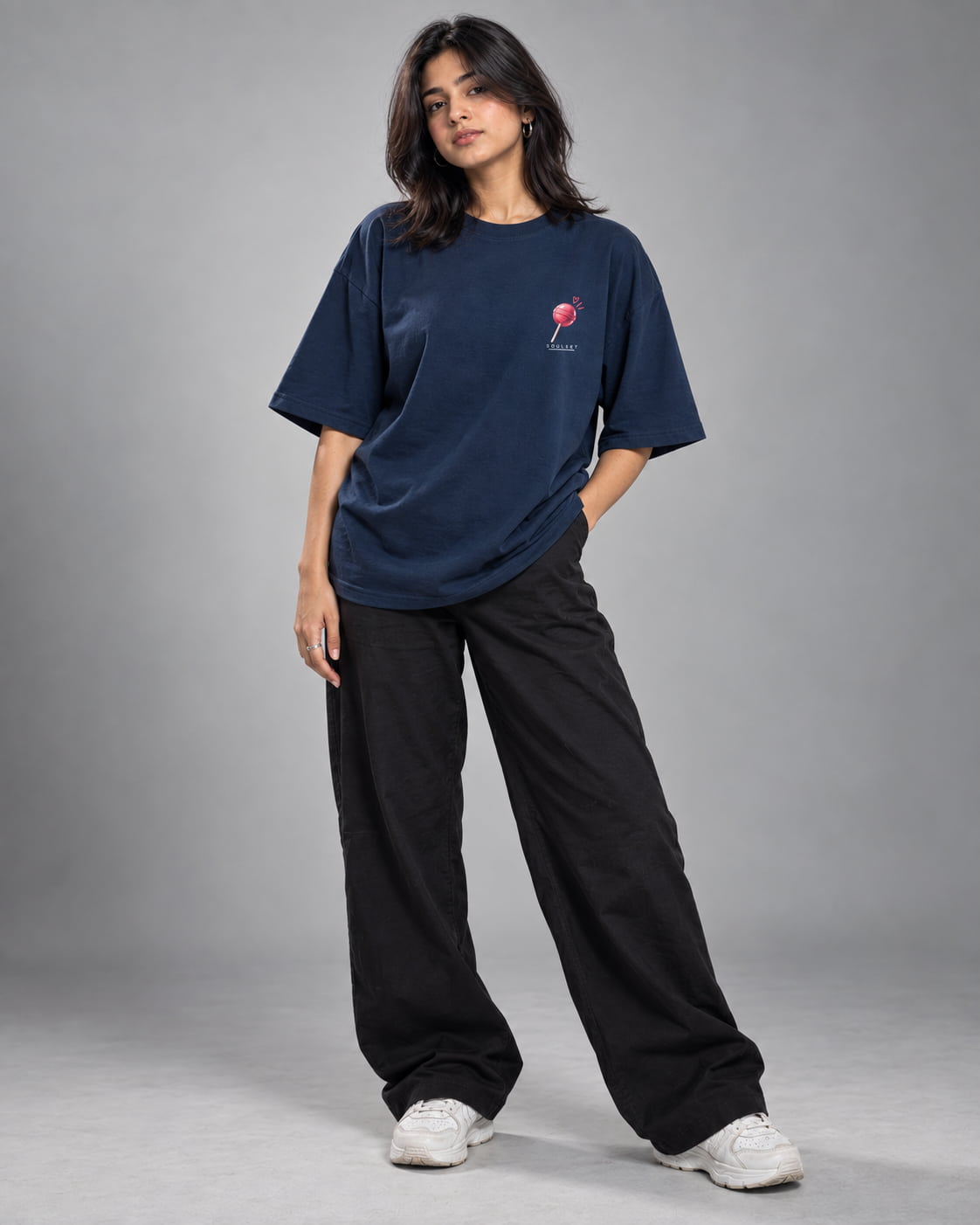 A woman modeling an oversized navy blue t-shirt with a small red and white graphic on the left chest, paired with loose black pants and white sneakers, against a solid gray studio background.