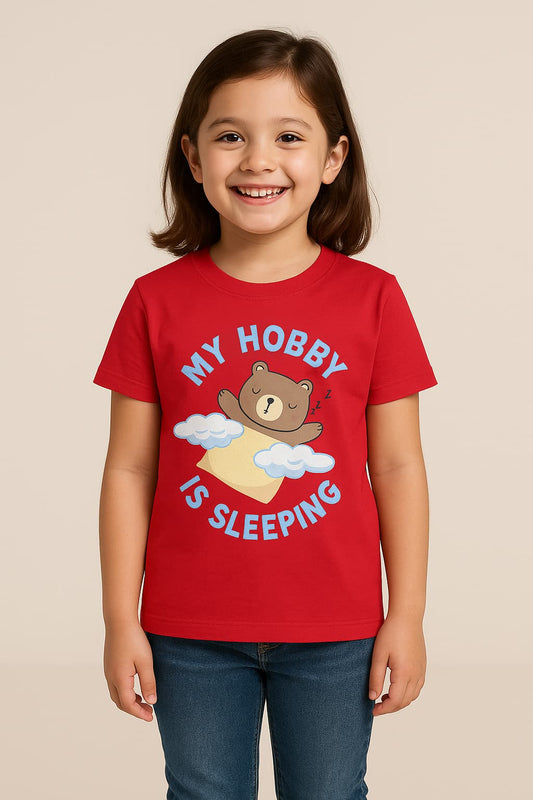 A cheerful young Indian girl with short dark hair smiles brightly in a studio with a neutral beige background. She is dressed in a red T-shirt featuring a sleeping bear graphic and the text “MY HOBBY IS SLEEPING,” along with casual blue jeans.