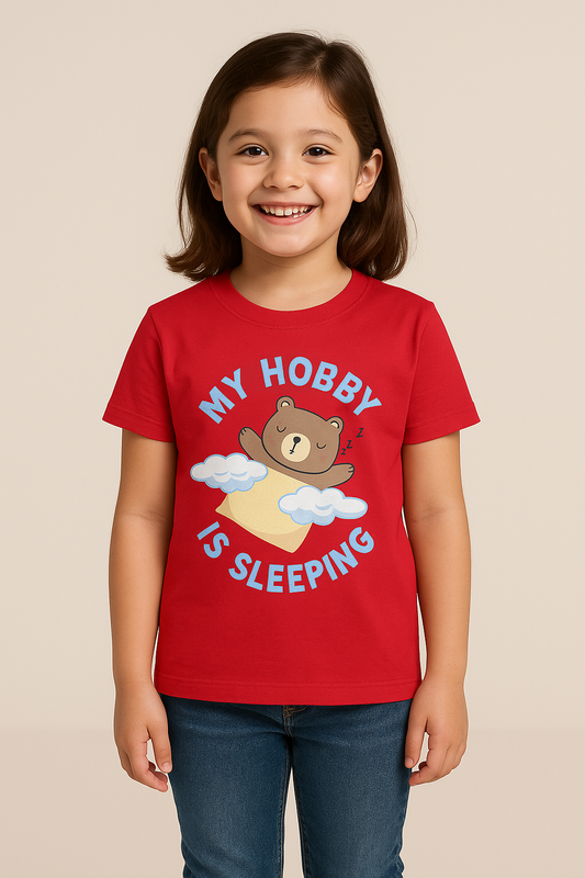 A cheerful young Indian girl with short dark hair smiles brightly in a studio with a neutral beige background. She is dressed in a red T-shirt featuring a sleeping bear graphic and the text “MY HOBBY IS SLEEPING,” along with casual blue jeans.