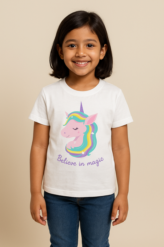 A digital photograph of a smiling 8-year-old Indian girl standing naturally against a clean, neutral studio background with soft lighting. She is wearing a white T-shirt featuring a colorful unicorn graphic with the text “Believe in magic,” paired with casual denim jeans. The photo is styled for an e-commerce clothing catalog, with the T-shirt as the main focus.