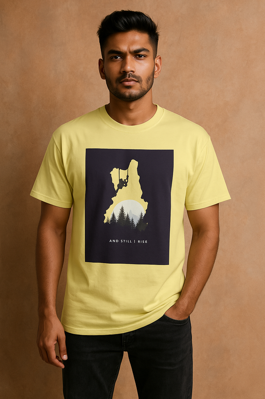A young South Asian male model stands confidently wearing a yellow oversized T-shirt with a bold adventure-themed print showing a climber silhouette, forest, and sun with the words “And Still I Rise.” He poses stylishly against an aesthetic background for an e-commerce catalog.