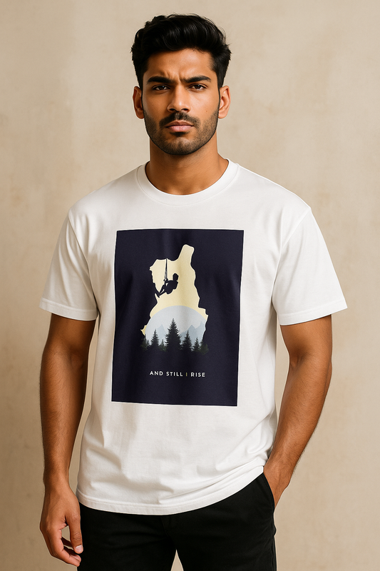 young South Asian male model in a relaxed pose wears a white oversized T-shirt featuring a climber silhouette with forest and sun design and the words “And Still I Rise.” The model looks bold and fashionable, styled for an online store photoshoot with a clean, modern background.