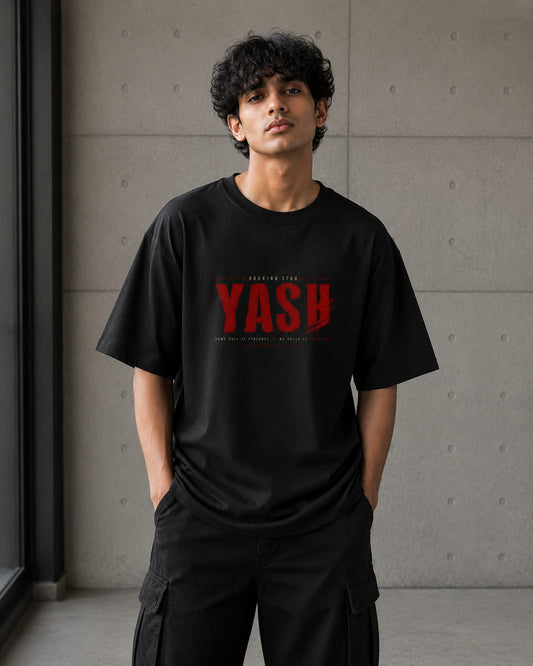 A man wearing a black crewneck t-shirt with a red distressed graphic reading 'YASH' on the chest, paired with black cargo pants, standing against a textured gray concrete wall.