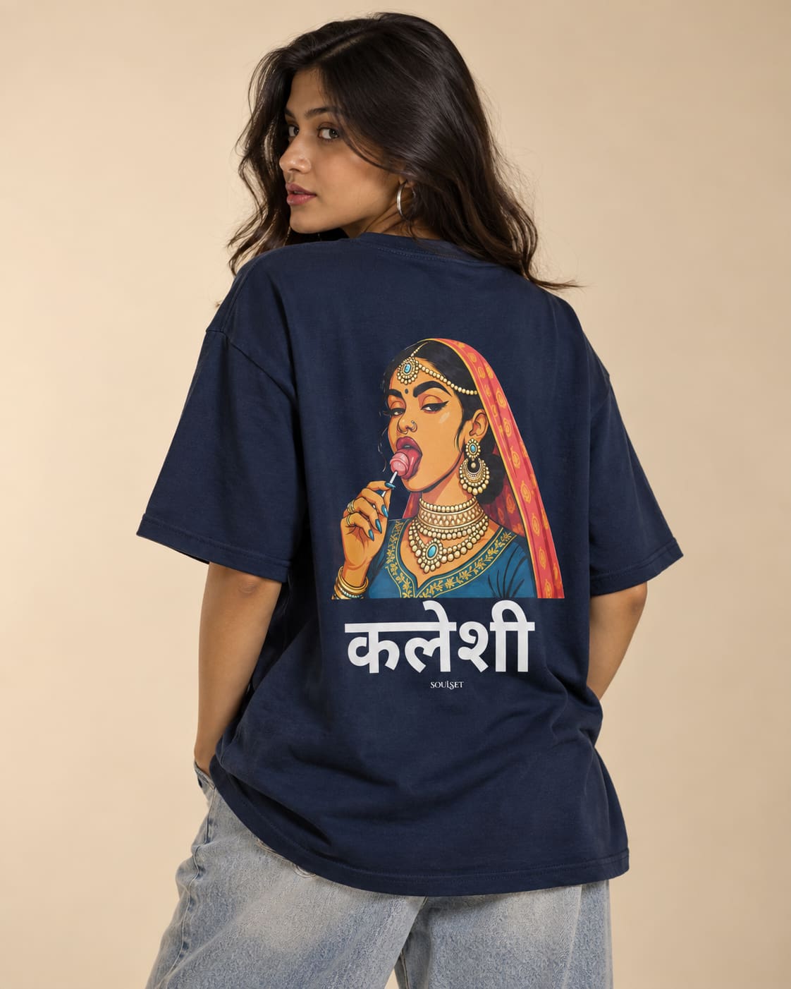 A woman wearing a navy blue t-shirt with a large graphic print of a traditional Indian woman and the text 'Kaleesh' on the back, paired with light wash jeans, set against a solid beige background.