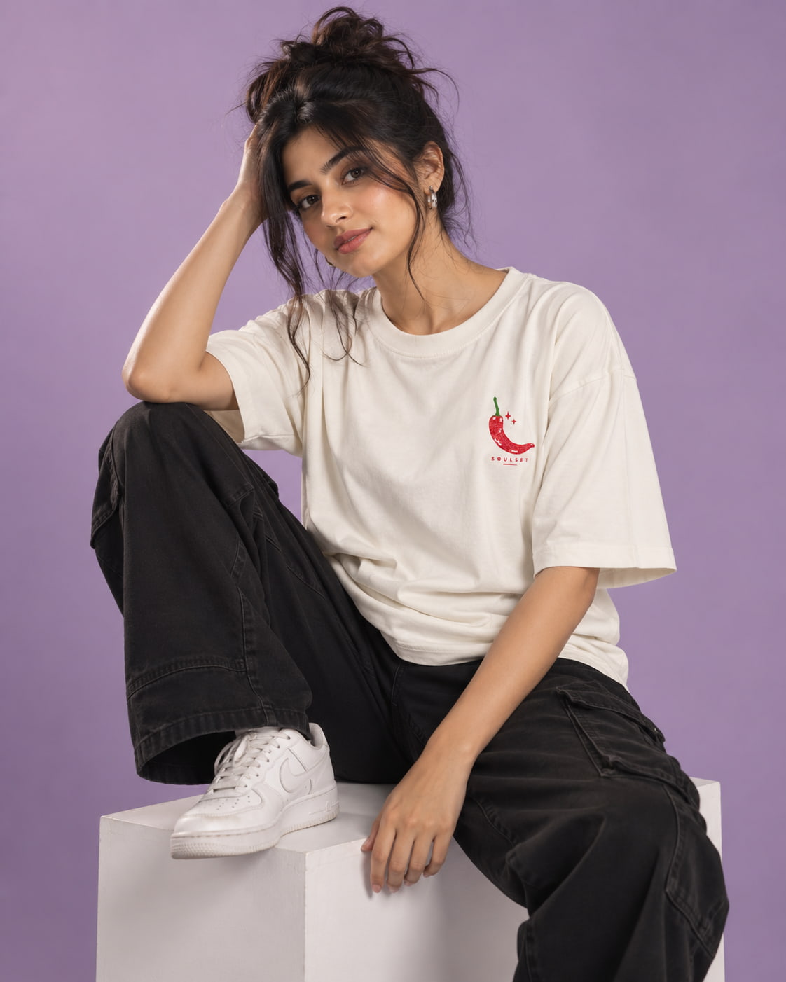 A woman models a white crewneck t-shirt featuring a small red chili pepper graphic on the left chest with the text 'CHILL OUT'. She is wearing black cargo pants and white sneakers, sitting on a white block against a solid purple background.