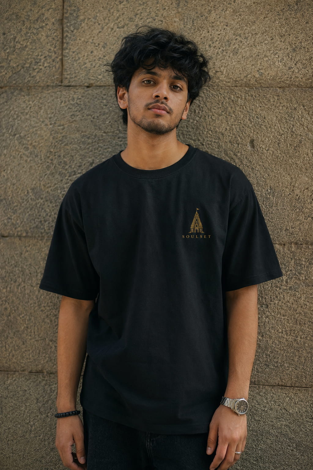 A man wearing a black crewneck t-shirt with a small gold graphic logo on the left chest, standing against a textured stone wall.